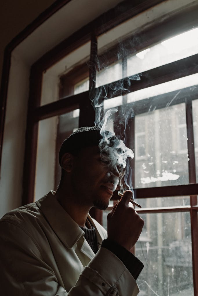 A man smoking by a window indoors, with natural light and smoke surrounding him.